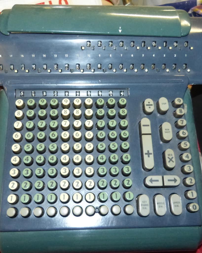The keyboard of an old blue-grey mechanical calculating machine photographed square on, ten columns of round number keys running from nine at the top down to a row of twos cut off along the bottom edge, the columns alternating in blocks of cream and pale green so about half the keys are green, a narrow strip of small numbered windows along the top edge, a right hand block of larger cream keys carrying a division sign, a STOP key, a short minus key above a long plus key, a NON SHIFT key and a key marked NEG with a cross on it, two more keys below them marked with arrows pointing left and right, and a single column of cream number keys down the far right edge