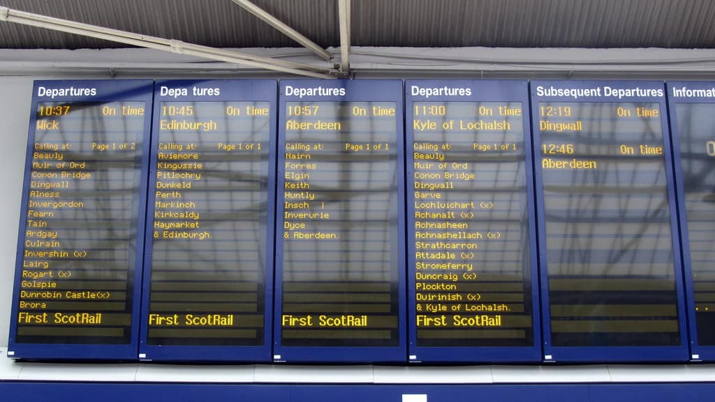 A row of six blue framed electronic departure screens mounted under a station roof, four of them headed Departures in white on blue with a yellow departure time and destination beneath, Wick, Edinburgh, Aberdeen and Kyle of Lochalsh, each of those four listing its calling points in small yellow type and carrying the operator name First ScotRail along the bottom, a fifth headed Subsequent Departures showing Dingwall and Aberdeen and carrying no operator name, a sixth cut off at the right edge headed Informat, and the words On time set beside every time on the board