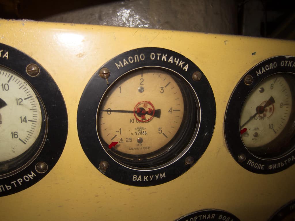 Three round pressure gauges set into a worn yellow instrument panel, the large central one carrying a long black needle resting at the low end of its scale and a short red index pointer below it, its black bezel stencilled in Cyrillic above the glass and again beneath it, a gauge at each side cut off by the frame with Cyrillic stencilled on their bezels in the same way, the left one showing a scale running from ten to sixteen and the right one carrying a black needle and a red pointer of its own