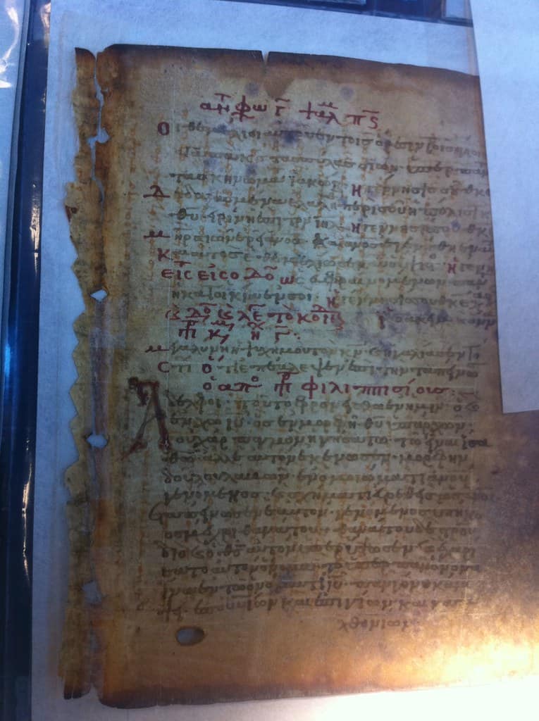 A stained parchment leaf in ochre and olive, closely covered in Greek script in dark brown ink with several lines and one large initial picked out in red, fainter marks visible between and behind the writing, the ragged left edge lit blue where a rod holds it against a pale mount