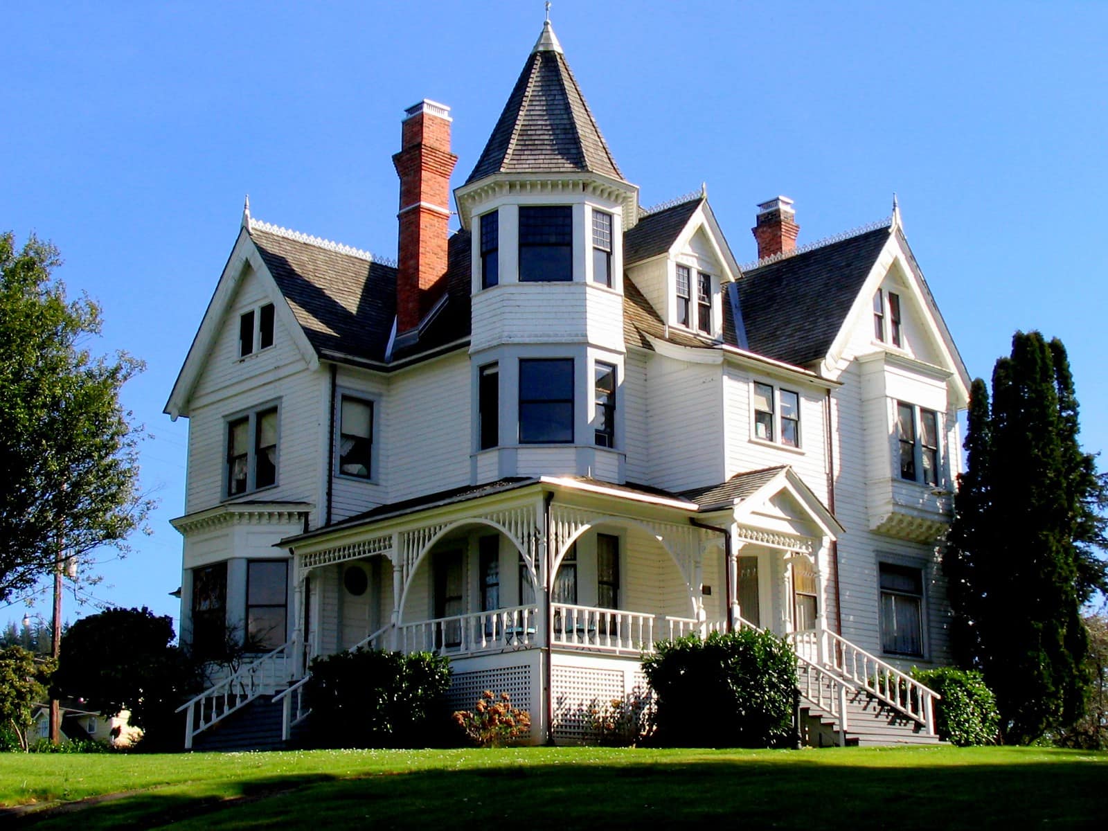 A large white Victorian house with a conical turret roof, two brick chimneys and a wraparound porch carried on turned posts with scrollwork brackets, photographed from below against a deep blue sky, with a tall dark cypress at the right edge