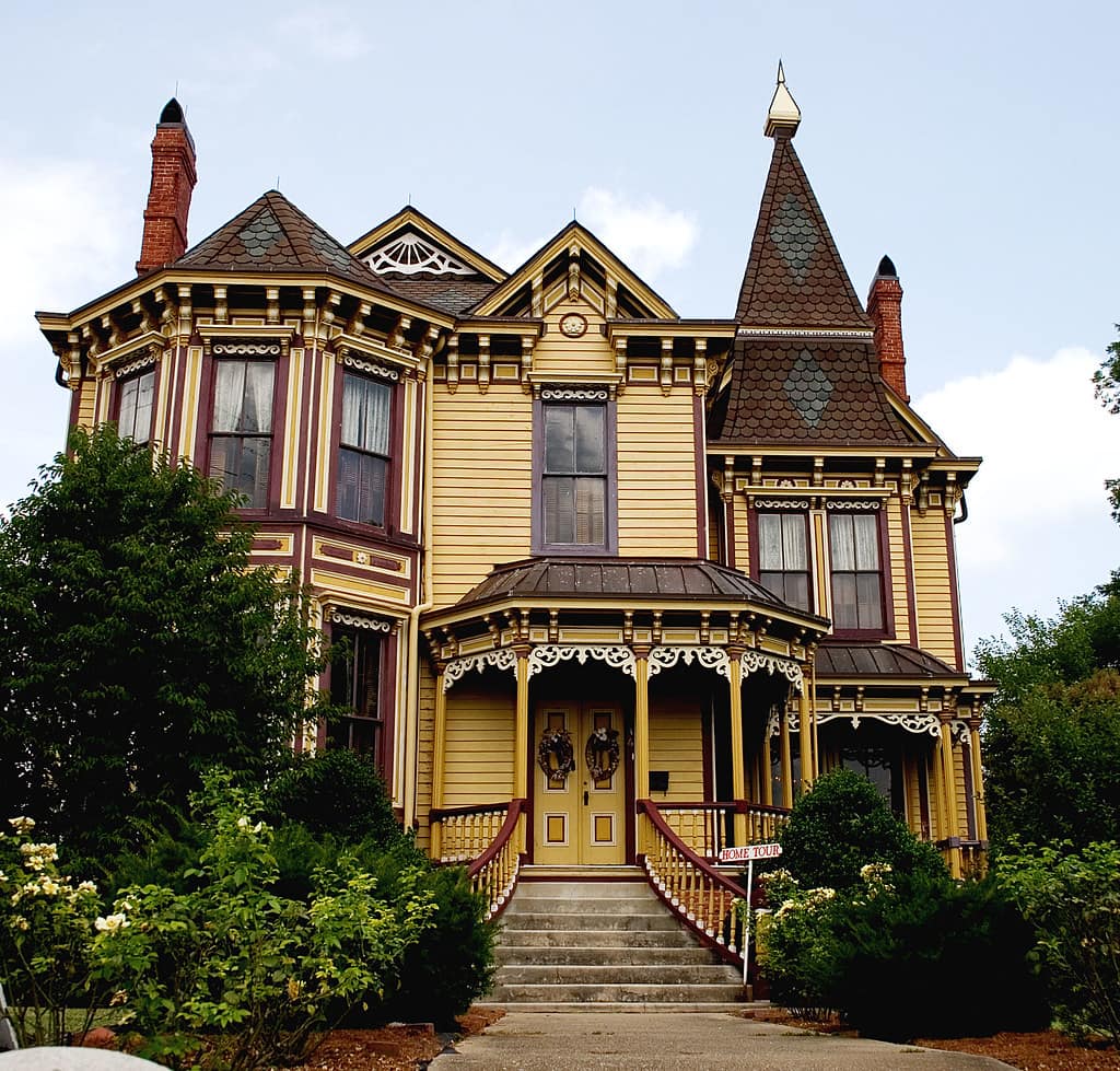 An ornate yellow and maroon Victorian house with a turret, a bracketed entrance porch under fretwork, curtains and blinds at most of the windows and two wreaths on the front door, photographed from the street under a white overcast sky with nobody in the frame and a white sign in red capitals standing at the foot of the steps, its wording cut off by the bottom of the frame