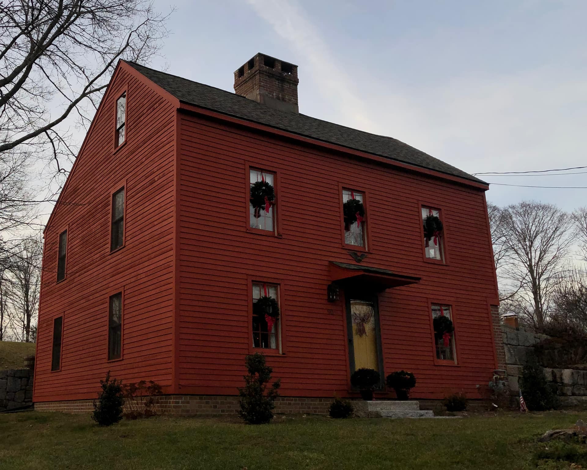 A red-painted saltbox house with a steep shingled roof, a brick chimney at the ridge and small-paned sash windows, the five windows across its front each hung with a dark evergreen wreath and a red bow and the door carrying a bow of its own, while the windows on the gable end at the left carry none, photographed from the road below under a pale grey winter sky