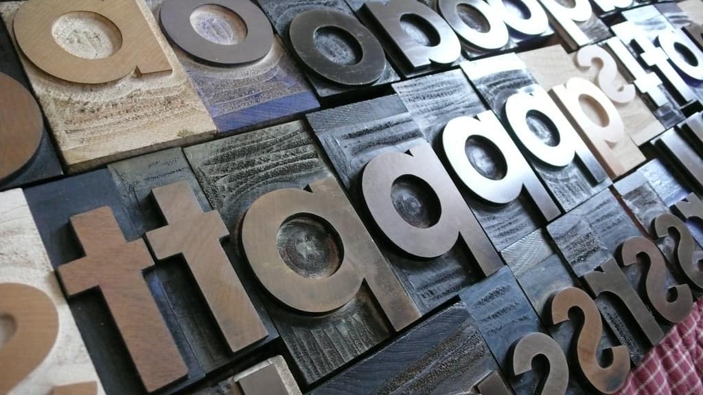 Large letterpress type blocks lying face up and packed tightly together, photographed at a low angle so the rows recede, a few of them pale bare wood and most of them darkened with ink, with lowercase t, a, o and p legible across the middle of the frame, a question mark among the blocks along the top edge, a row of figures at the right hand edge, and a fold of pink and white checked cloth at the bottom right corner
