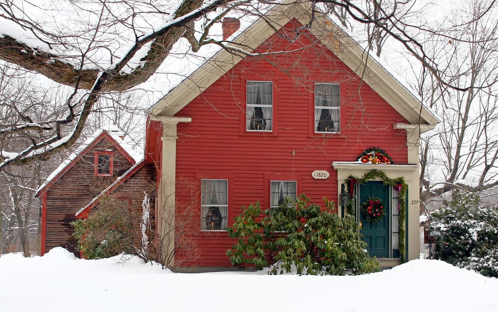 A red clapboard house in snow with cream window frames and electric candles in several of them, an oval plaque reading c 1820 on the wall and the number 279 painted beside a dark teal front door hung with a red berried wreath under a green garland, with a second, weathered red building standing behind it to the left and snow laden bare branches crossing the top of the frame