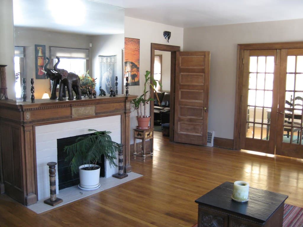 A living room with a wooden fireplace mantel carrying two large carved elephants and a row of turned candlesticks under a tall mirror, a fern in a white pot on the hearth in front of a black firebox whose surround is painted white, a five panel door standing open onto a further room with a window and a sofa in it, and glazed French doors at the right throwing sunlight across a polished wood floor