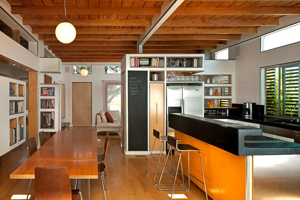 An open plan room with exposed timber ceiling beams, a long polished wooden dining table on the left and an orange fronted kitchen island with metal stools on the right, and in the centre a white cabinet whose front is a dark chalkboard panel carrying a handwritten list in pale green chalk reading milk, dog food, coffee, bread, cheese and soap